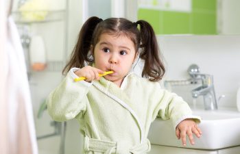 A young child with pigtails brushes her teeth in a bathroom while wearing a light green bathrobe.